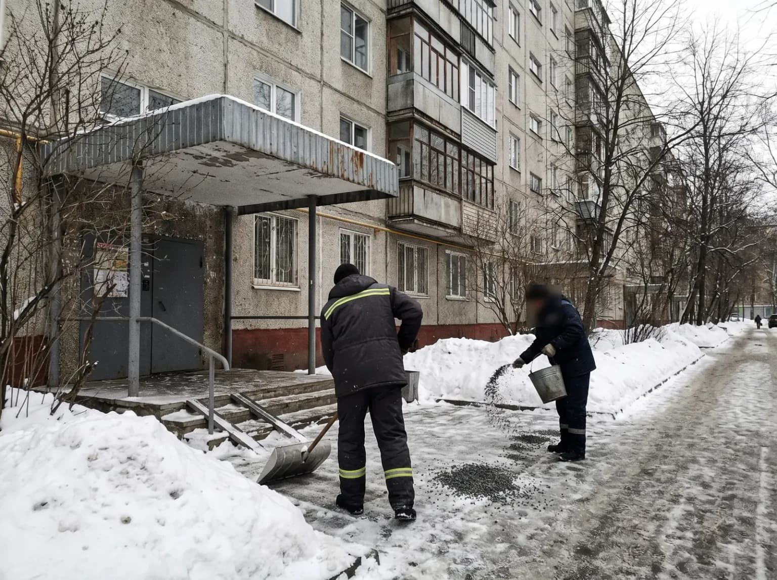 Рабочие чистят ступени подъезда и посыпают тротуар крошкой